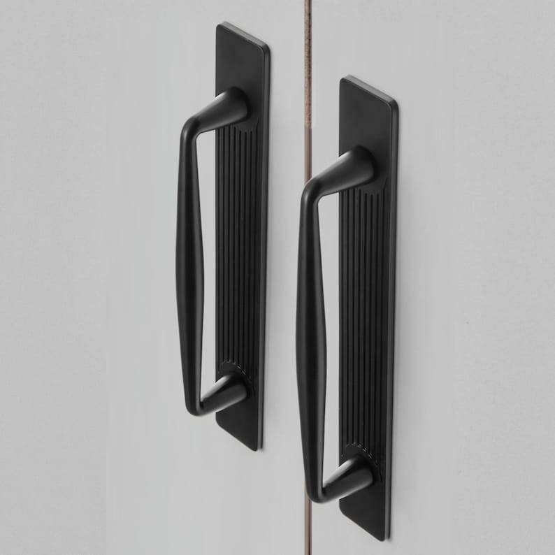 May include: Two black cabinet pulls with a rectangular backplate and a curved handle. The backplate has vertical grooves. The handles are mounted on a white surface.