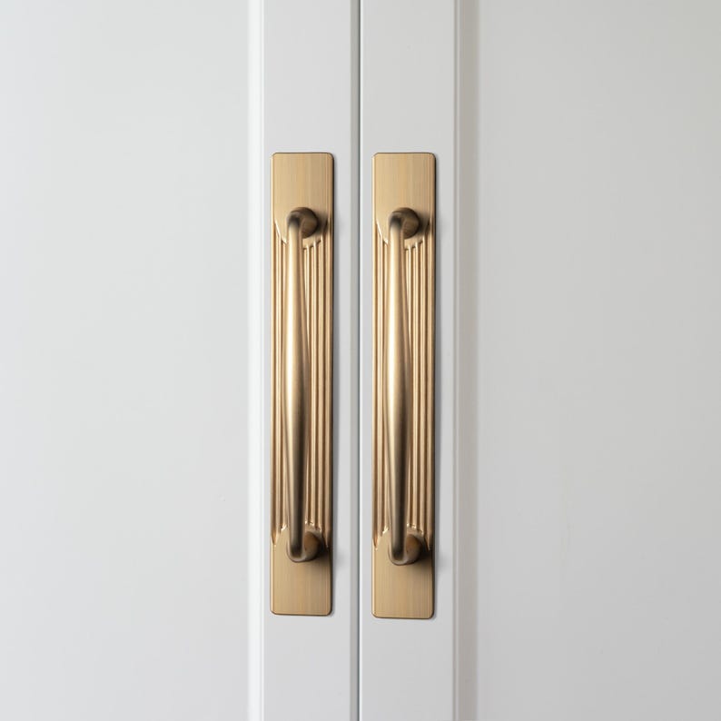 May include: Two gold-colored cabinet handles with a vertical ribbed design are mounted on a white cabinet door. The handles have a rectangular backplate and a curved, cylindrical grip. The hardware adds a touch of elegance to the cabinet.