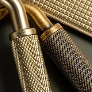 Op de afbeelding: Close-up van antieke messing hardware, met getextureerde handgrepen en een rechthoekige plaat. De handgrepen hebben diamantvormige grepen en het metaal heeft een warme, verouderde afwerking. De hardware is waarschijnlijk voor een kast of lade.