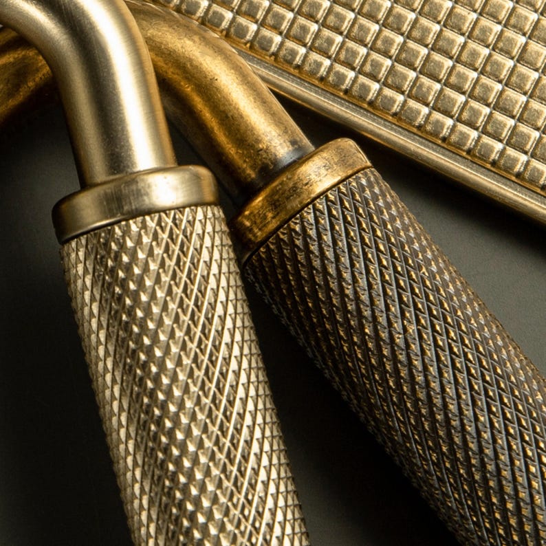 Op de afbeelding: Close-up van antieke messing hardware, met getextureerde handgrepen en een rechthoekige plaat. De handgrepen hebben diamantvormige grepen en het metaal heeft een warme, verouderde afwerking. De hardware is waarschijnlijk voor een kast of lade.
