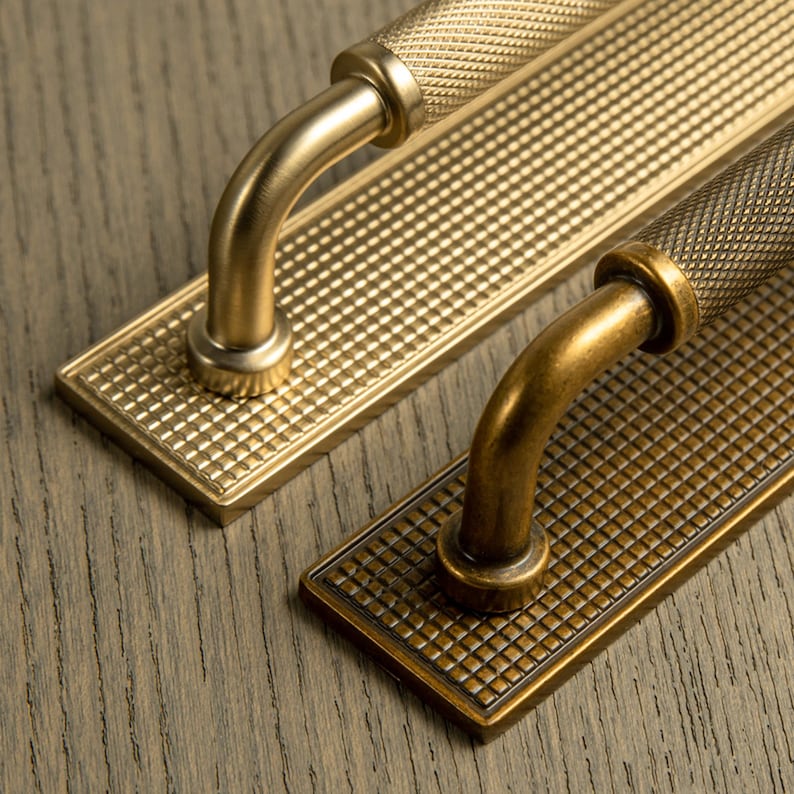 Op de afbeelding: Close-up van twee messing deurgrepen. Elk heeft een getextureerd, rasterachtig patroon op de rechthoekige basis en de handgreep. De handgrepen zijn gebogen. E&eacute;n is lichter messing, de andere donkerder, antiek messing.