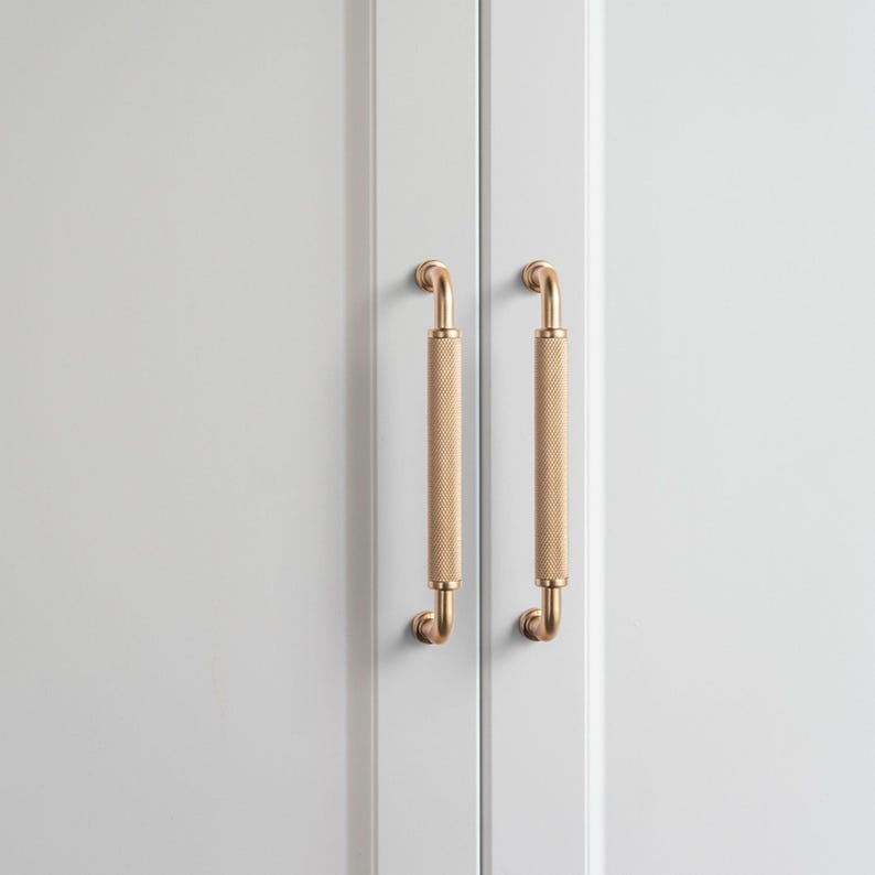 Op de afbeelding: Twee goudkleurige kastgrepen met een getextureerde, cilindrische grip en gebogen uiteinden zijn gemonteerd op een lichtgrijze kastdeur. De grepen zijn verticaal uitgelijnd en bieden een modern en elegant ontwerp.