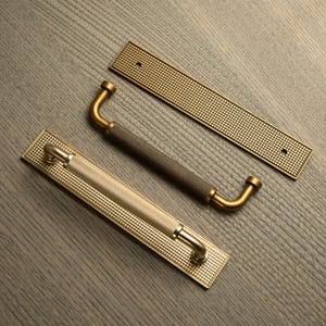 Op de afbeelding: Drie decoratieve deurgrepen in messing en donkerbruin. De grepen hebben een rechthoekige achterplaat met een rasterpatroon. De handgrepen zijn gebogen en getextureerd. De hardware is ontworpen voor kasten en deuren.