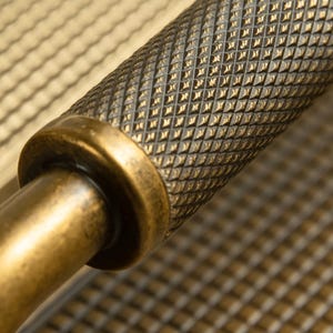 Op de afbeelding: Close-up van een antieke messing deurklink. De klink heeft een diamantpatroon grip en een cirkelvormige basis. Het messing heeft een verweerde, verouderde uitstraling, met kleurvariaties van goud tot donkerbruin. De achtergrond is een getextureerd oppervlak.