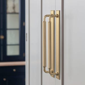 Op de afbeelding: Close-up van twee goudkleurige kastgrepen. Elke handgreep heeft een getextureerde, cilindrische grip en is gemonteerd op een rechthoekige achterplaat met verticale groeven. De handgrepen zijn bevestigd aan een lichtgrijze kastdeur.