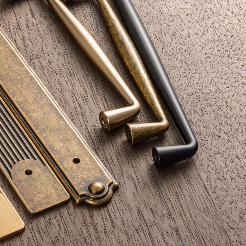 May include: Close-up of various cabinet hardware in antique brass, gold, and black finishes. The selection includes pull handles and rectangular backplates, showcasing different styles and designs for home decor and renovation projects.