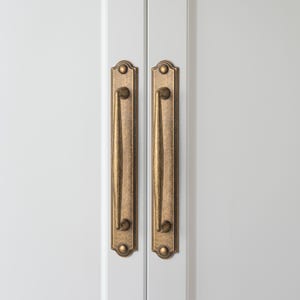 May include: Two antique brass cabinet pulls with a vertical design. The pulls are mounted on a white cabinet door. Each pull has a rectangular backplate with decorative detailing and a curved handle.