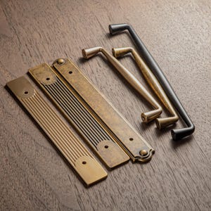 May include: A collection of antique-style cabinet hardware, including brass and black metal handles and pulls. The hardware features various designs, including bar pulls and handles with decorative details. The items are arranged on a dark wood surface.