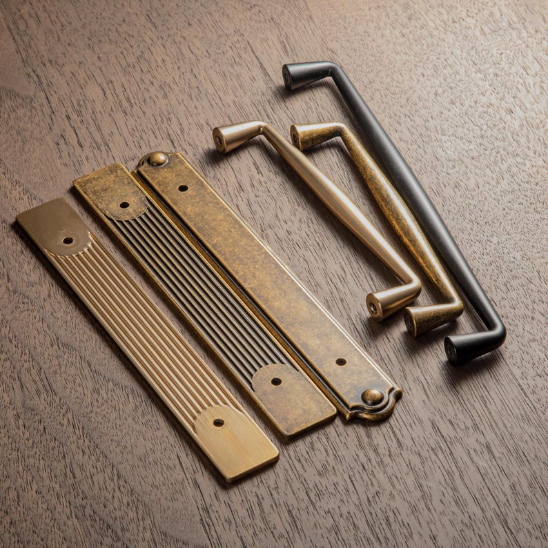 May include: A collection of antique-style cabinet hardware, including brass and black metal handles and pulls. The hardware features various designs, including bar pulls and handles with decorative details. The items are arranged on a dark wood surface.