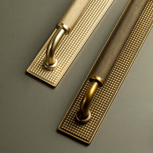 Op de afbeelding: Twee messing deurklinken met een textuur, rasterachtig patroon. De handgrepen hebben een gebogen ontwerp en zijn gemonteerd op rechthoekige achterplaten. De afwerking is een warme, antieke goudkleur, geschikt voor interieurdecoratie.