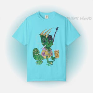 May include: Light blue t-shirt featuring a cartoon chameleon with a crown, smoking a pipe, and holding a glass of what appears to be gold nuggets. The chameleon is green, yellow, and purple. The text "MUTINY FARMS" is printed on the shirt.