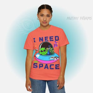 Op de afbeelding: Koraalrood T-shirt met de tekst "I NEED SPACE" in blauwe blokletters. De afbeelding toont een groene alien in een ruimteschip met andere kleurrijke wezens. Het shirt is een ronde hals.