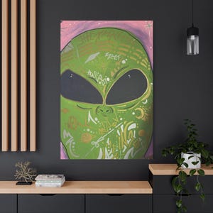 Op de afbeelding: Portret van een groene alien op canvas, met grote zwarte ogen en gouden en zilveren graffiti-achtige ontwerpen. De achtergrond is zachtroze met witte sterren. Het kunstwerk wordt aan een muur getoond.