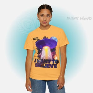 Op de afbeelding: Mosterdgeel T-shirt met een paars UFO-ontwerp, een heldere lichtstraal en de tekst "I WANT TO BELIEVE". Het shirt heeft ook de tekst "MUTINY FARMS" en "NEW FARMS".