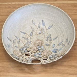 May include: A handcrafted ceramic bowl with a floral design. The bowl features a light beige color with embossed flowers in shades of blue and pink. The rim has a series of decorative cut-out holes. The bowl is placed on a wooden surface.