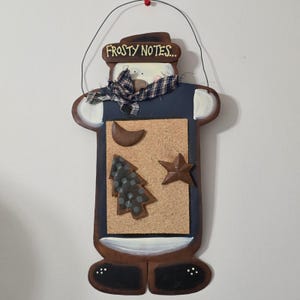 May include: A snowman-shaped corkboard with the text "Frosty Notes...". The board features a brown frame, a blue body, and a plaid scarf. Decorative elements include a tree, star, and crescent moon.