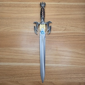 May include: A decorative sword with a silver blade and ornate hilt. The hilt features a gold and silver crossguard with intricate detailing. The handle is brown and silver, and the pommel has a crown-like design. The sword is displayed on a wooden surface.