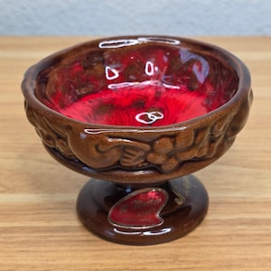 May include: A small ceramic pedestal bowl featuring a glossy brown exterior with a relief pattern of grapes and leaves. The interior is glazed in a vibrant, deep red with dark brown accents. A red sticker on the base displays the text In Master.