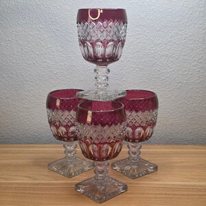 May include: A set of four wine glasses featuring a deep cranberry red upper bowl with a diamond-cut pattern and a clear glass stem and square base. The glasses are arranged with one glass stacked on top of three others in a cluster formation.