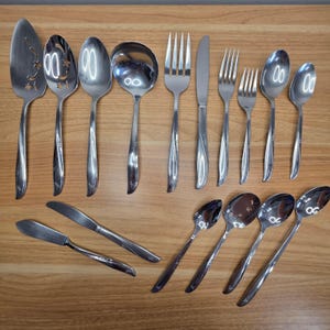May include: A collection of stainless steel silverware, including spoons, forks, knives, and serving utensils. The silverware has a shiny finish and a decorative starburst design on the handles. The pieces are arranged on a light brown wooden surface.