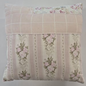 May include: Square decorative pillow with a patchwork design. The pillow features a light pink grid pattern, floral rose print, and a floral rose pattern. The pillow is a square shape and appears to be made of fabric.
