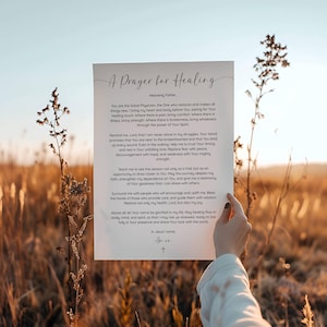 May include: A printed paper with the text "A Prayer for Healing" in elegant script. The prayer is written in a classic font, with a cross at the bottom. The paper is held against a blurred background of a field.