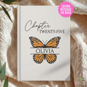 May include: A white hardcover book featuring the black text Chapter TWENTY-FIVE and a monarch butterfly graphic with the name OLIVIA centered across the wings. A pink circular badge in the top right corner displays the text LOVING MESSAGE ON BACK.