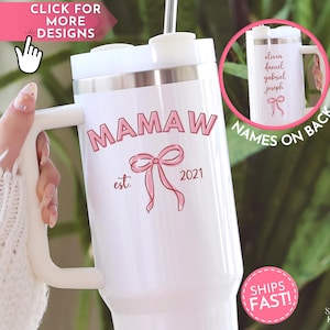 May include: White insulated tumbler with a handle, featuring the text "MAMAW est. 2021" in pink, along with a pink bow graphic. The back of the tumbler shows names. The tumbler has a silver band and a white lid.