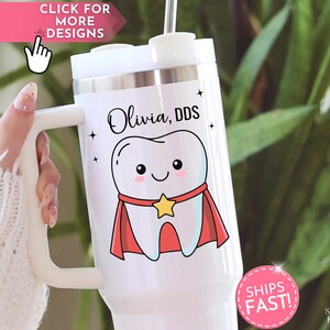 May include: White tumbler with a handle, featuring a cartoon tooth wearing a cape and a star. The tumbler has the name "Olivia, DDS" and small star accents. A pink sticker reads "SHIPS FAST!"