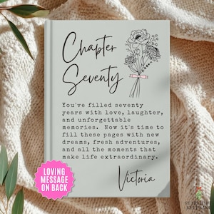 May include: A light gray hardcover book featuring the text Chapter Seventy and a floral line drawing. Below, the text reads: You've filled seventy years with love, laughter, and unforgettable memories. Now it's time to fill these pages with new dreams, fresh adventures, and all the moments that make life extraordinary. The name Victoria is printed at the bottom, and a pink starburst graphic states: LOVING MESSAGE ON BACK.