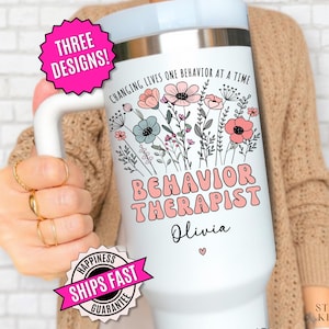 May include: A white stainless steel tumbler with a handle featuring a floral graphic, the text "CHANGING LIVES ONE BEHAVIOR AT A TIME", "BEHAVIOR THERAPIST", and the name "Olivia" in black and pink script. A pink starburst graphic reads "THREE DESIGNS!"