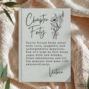May include: A light gray hardcover book with the text Chapter Forty and a floral illustration in black ink. The text reads: You've filled forty years with love, laughter, and unforgettable memories. Now it's time to fill these pages with new dreams, fresh adventures, and all the moments that make life extraordinary. The name Victoria is printed at the bottom.