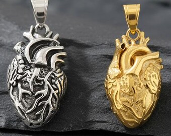 Può includere: Due ciondoli a forma di cuore anatomico. Uno è color argento, l'altro dorato. Entrambi i ciondoli presentano vene e arterie dettagliate e hanno un'asola per una catena. Sono esposti su una superficie strutturata grigio scuro.