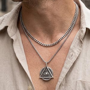 Könnte beinhalten: Ein mehrlagiges silberfarbenes Halsketten-Set bestehend aus einer kürzeren Panzerkette und einer längeren Kette mit einem runden Anhänger. Der Anhänger zeigt ein dreieckiges Design mit einem Auge und strahlenförmigen Linien.