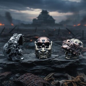 May include: Three detailed skull-shaped rings in silver, bronze, and copper tones rest on a dark, rocky surface. The rings feature intricate designs, with a backdrop of a castle and a fiery, war-torn landscape.