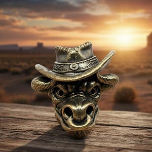 May include: A detailed bronze-colored bull head sculpture wearing a cowboy hat. The sculpture has intricate details and a ring in its nose. The background shows a desert landscape with a sunset and a campfire.