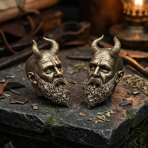 May include: Two bronze sculptures of horned, bearded faces. The sculptures have detailed facial features and curved horns. They are placed on a textured, grey stone surface, suggesting a thematic or decorative item.