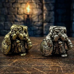May include: Two metal dwarf figurines, one gold and one silver, are presented on a wooden surface. Each dwarf holds a shield and is detailed with a beard and armour. The background features a stone wall and a warm light source.