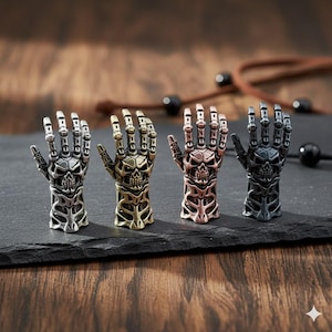 May include: Four metal skeleton hand sculptures in silver, gold, copper, and black. Each hand features a skull design and intricate skeletal detailing. The hands are displayed on a dark, textured surface.
