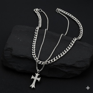 May include: A silver layered necklace with a cross pendant. The necklace features two chains: a thicker curb chain and a thinner chain. The cross pendant is adorned with small, sparkling embellishments. The jewelry is displayed against a dark, textured background.