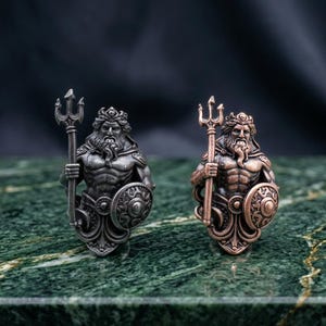 May include: Two metal figurines depicting a bearded figure with a crown, holding a trident in one hand and a circular shield in the other. One figurine has a dark silver finish, while the other has a warm copper finish, both showing detailed relief.