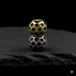 Könnte beinhalten: Zwei Metallperlen, eine goldfarbene und eine silberfarbene, übereinander gestapelt. Jede Perle ist kugelförmig und weist zahlreiche, gleichmäßig verteilte kreisförmige Löcher auf. Die goldfarbene Perle liegt auf der silberfarbenen, vor dunklem Hintergrund.