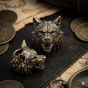 Puede incluir: Dos anillos con cabeza de lobo en bronce con detalles de pelaje y bocas abiertas. Los anillos están sobre una superficie de cuero con monedas antiguas y una brújula de fondo. Un accesorio único.