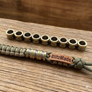 May include: A close-up shot of a paracord lanyard with brass beads. The lanyard is olive green and tan, with a series of brass beads and a leather tag that reads "BlitzMade". The beads are cylindrical with a hole through the center.