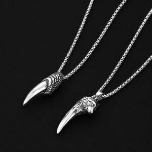 May include: Two silver-colored pendant necklaces. One features a claw design, the other a wolf head. Both pendants hang from a silver chain. The background is a solid black.
