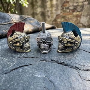 May include: Three metallic skull figurines featuring detailed bone textures and rectangular sunglasses. The left skull has a red mohawk, the center skull is silver with a plain crest, and the right skull has a blue mohawk. Each skull is cast in metal.