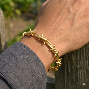 May include: A gold-colored barbed wire bracelet. The bracelet is wrapped around a wrist, with the sharp points of the wire facing outward. The bracelet is made of a shiny, metallic material.