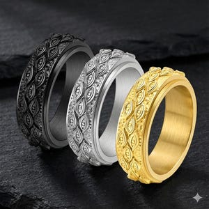 May include: Three metal spinner rings in black, silver, and gold. Each ring features a textured band with an eye-like pattern. The rings are displayed on a dark, textured surface, showcasing their design and metallic finishes.