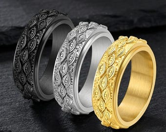 May include: Three metal spinner rings in black, silver, and gold. Each ring features a textured band with an eye-like pattern. The rings are displayed on a dark, textured surface, showcasing their design and metallic finishes.