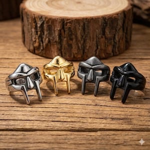 May include: Four rings in silver, gold, gunmetal, and black, each featuring a stylized mask design. The rings are displayed on a wooden surface with a wood slice in the background. The rings are likely made of metal.